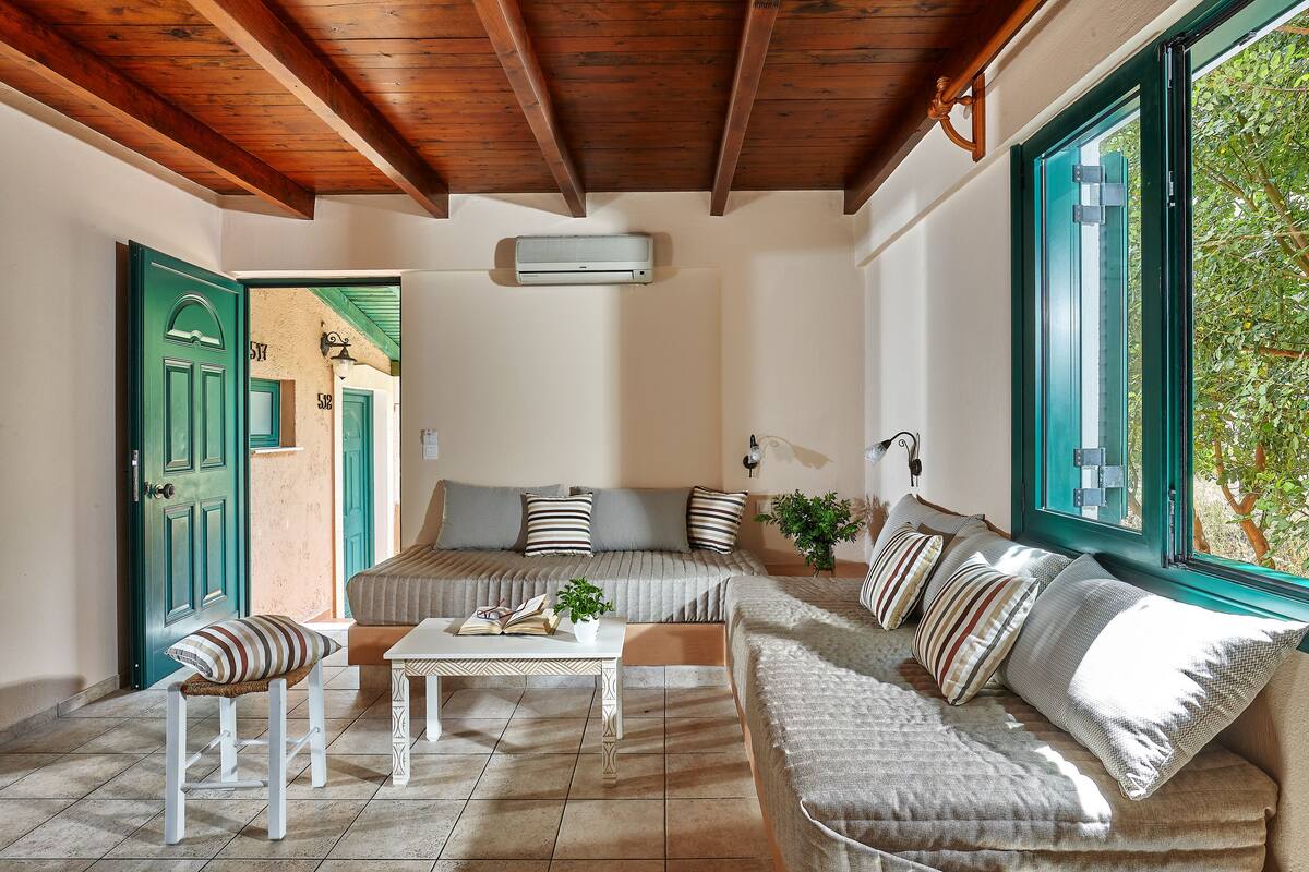 A cozy living room with a sofa, a small table, and a bench. There is a window with a view of greenery outside.