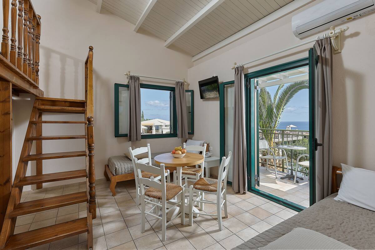 A room with a wooden staircase, a round dining table with chairs, a bed, and a large window with curtains.