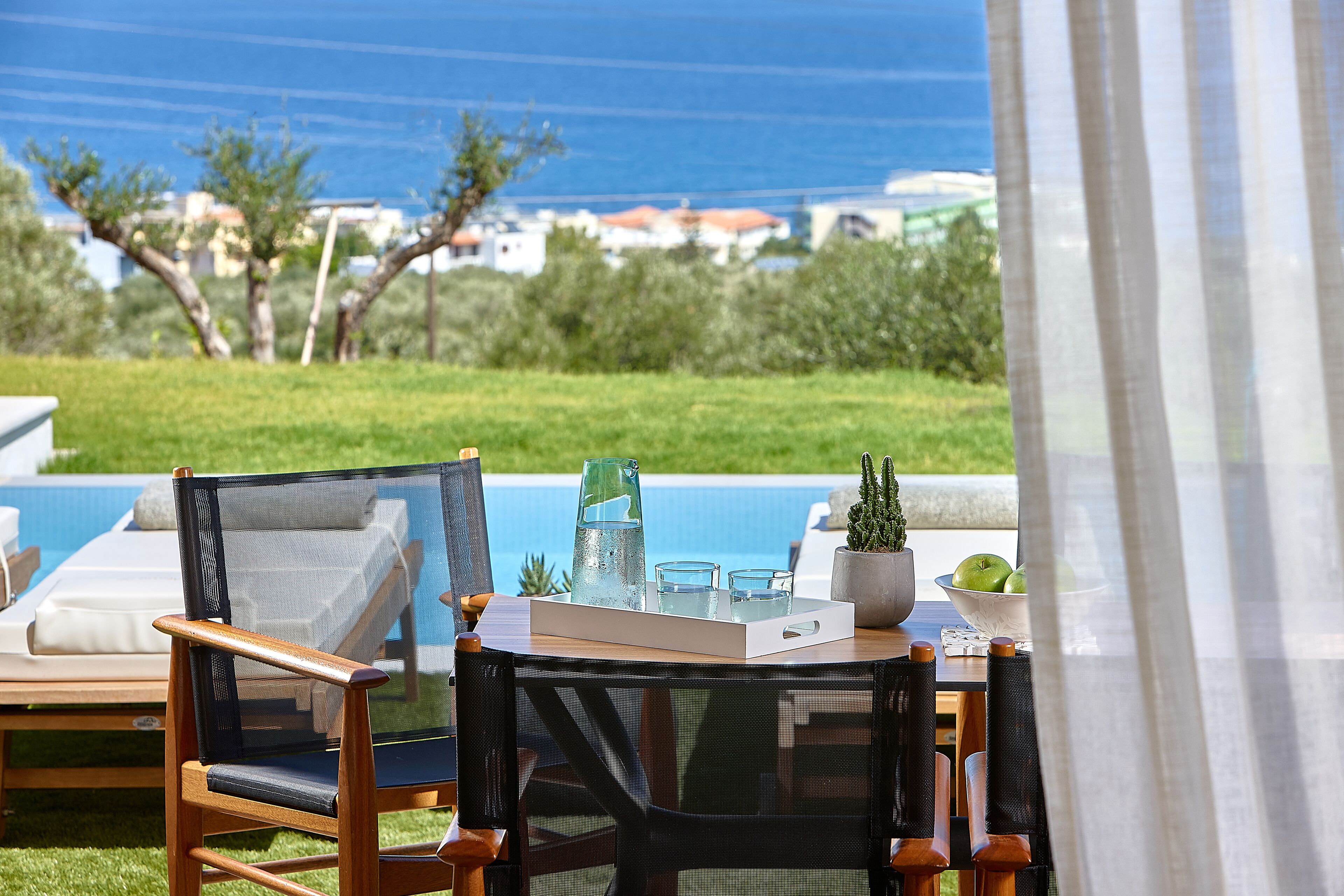 Uma área de estar à beira da piscina com mesa, cadeiras e um tabuleiro com bebidas e jarra.