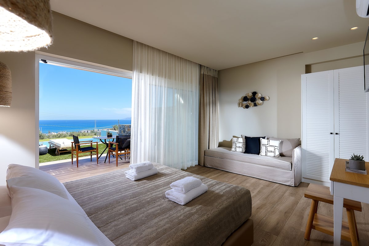 A modern hotel room with a large bed, a sofa, a dining area, and a view of the ocean.