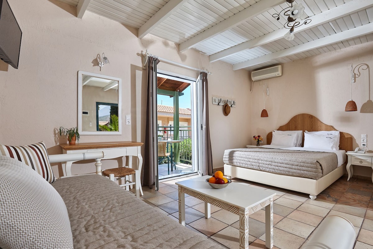 A bedroom with a bed, a desk, a chair, a small table with fruit, and a balcony with a view.