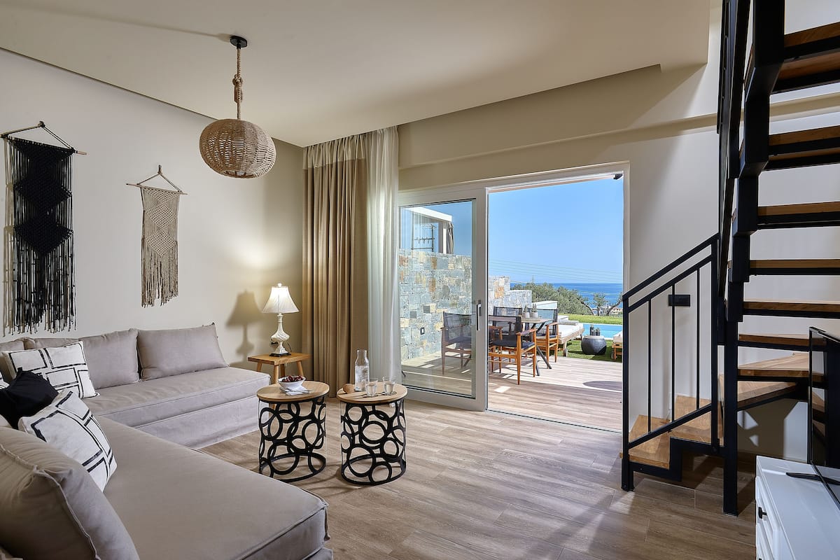 A modern living room with a sofa, two side tables, a hanging light fixture, and a view of a balcony with outdoor furniture.