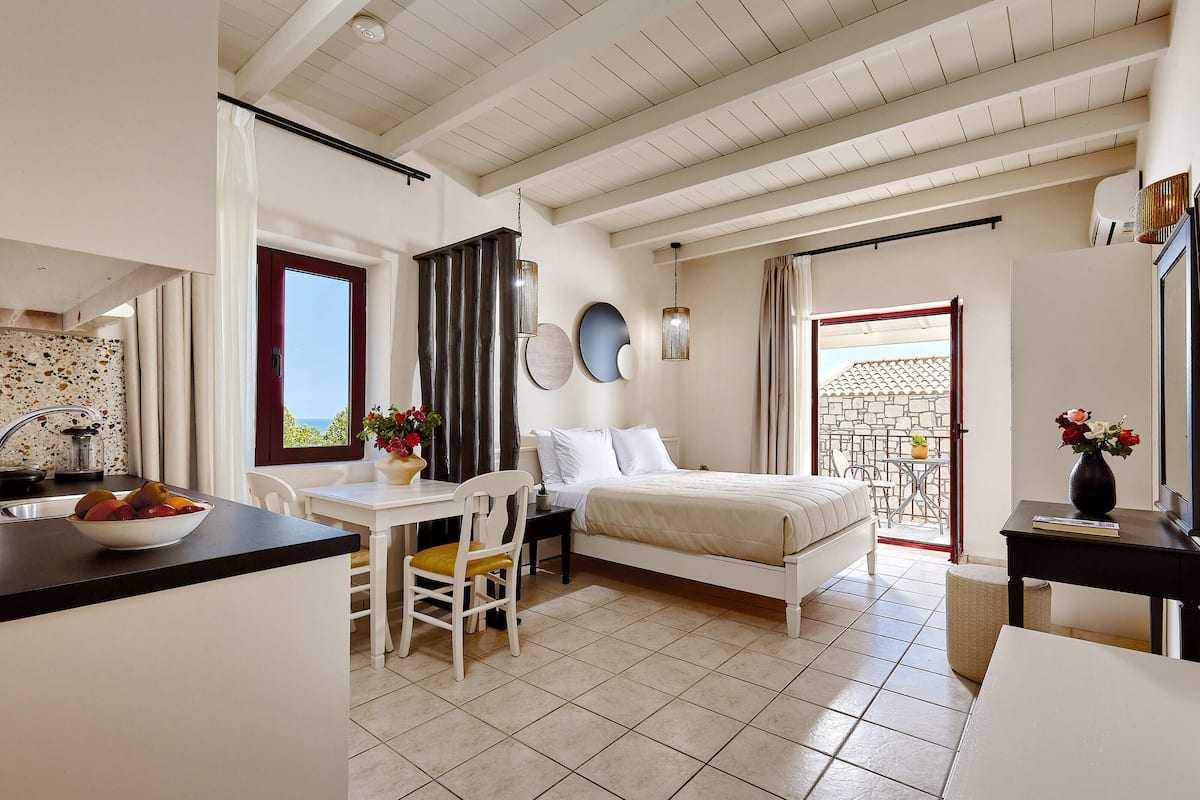 A bedroom with a bed, a desk, and a chair. There is a window with curtains, a balcony with a table and chairs, and a view of the sea.