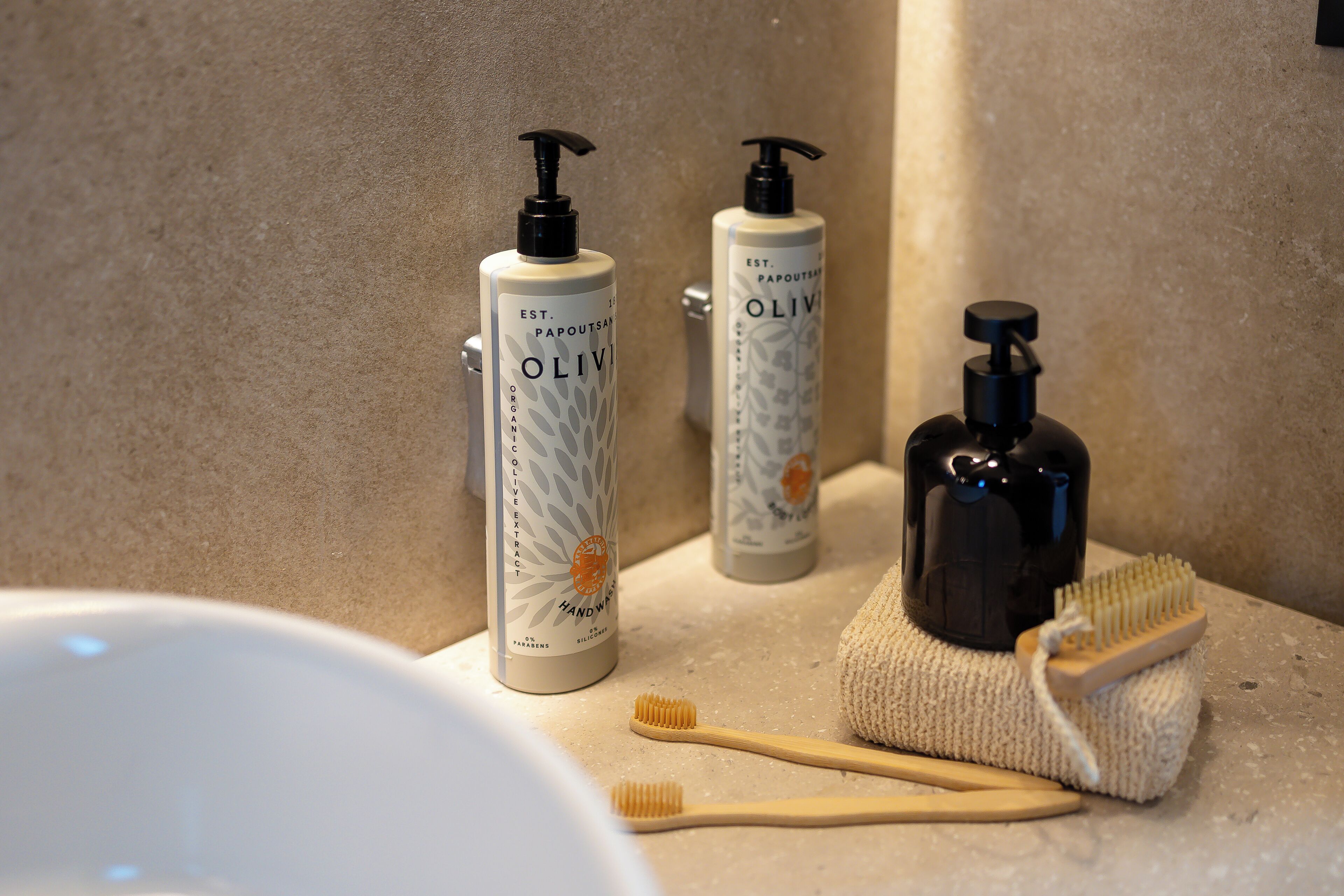 A bathroom with two bottles of olive oil, a black soap dispenser, and two wooden toothbrushes on a beige countertop.