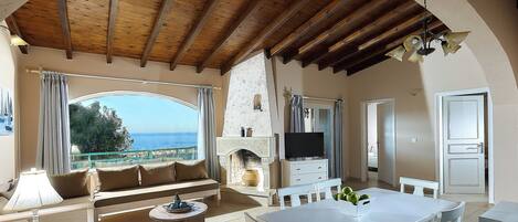 A spacious living area with a wooden ceiling, a fireplace, a television, and a view of the sea.