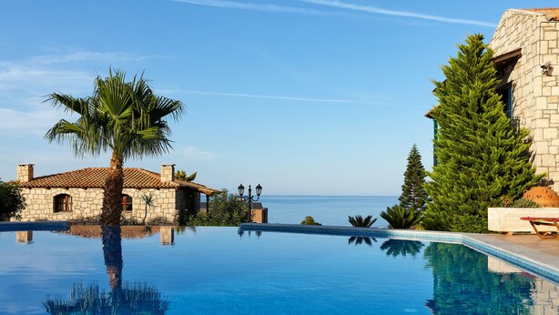 A clear blue infinity pool with a view of the sea, surrounded by stone buildings and palm trees. - Marni Village (Hersonissos)