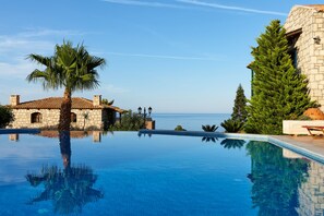 Una piscina infinita azul y cristalina con vistas al mar, rodeada de edificios de piedra y palmeras.