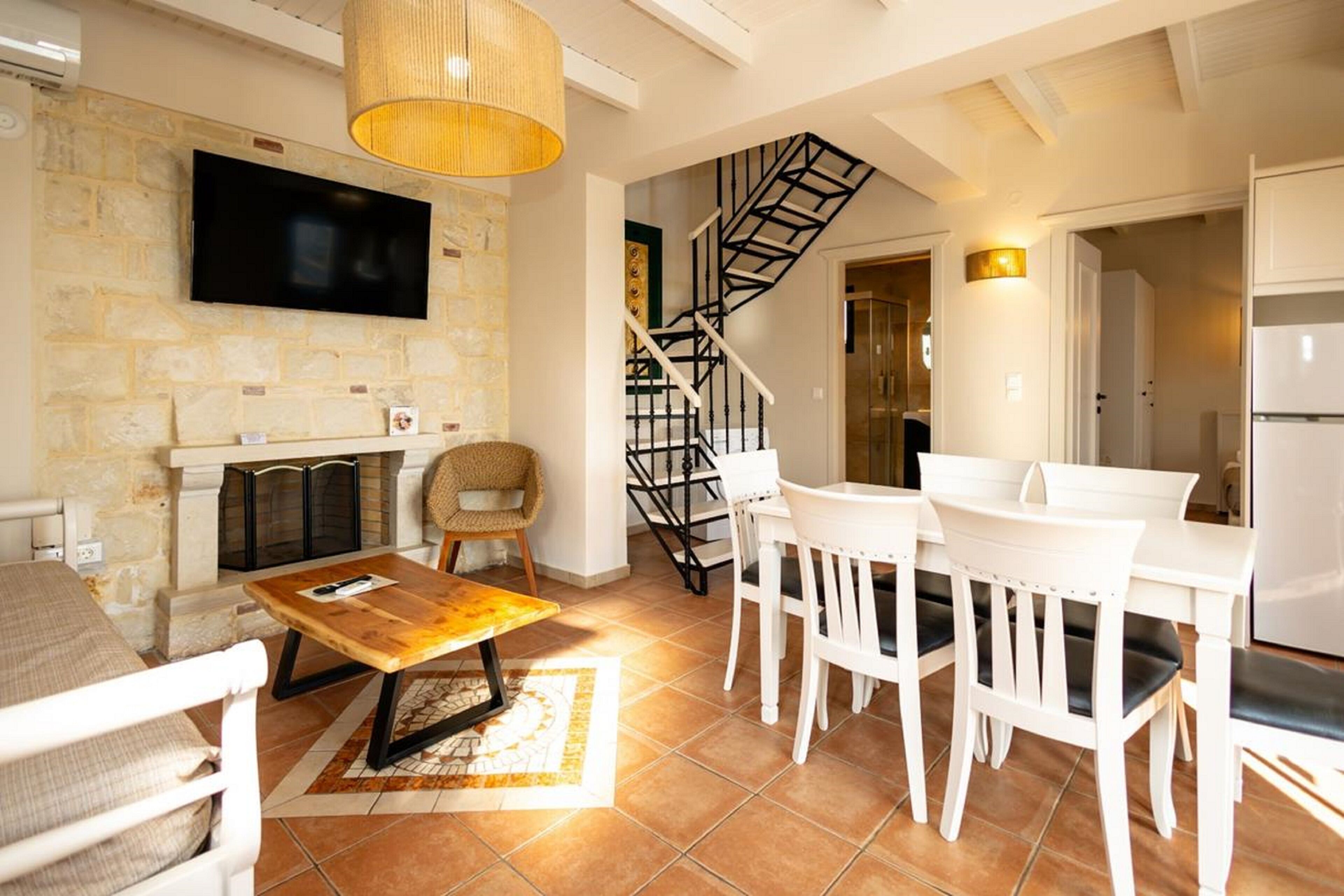 A living room with a fireplace, a TV mounted on a stone wall, a wooden coffee table, white furniture, and a spiral staircase.