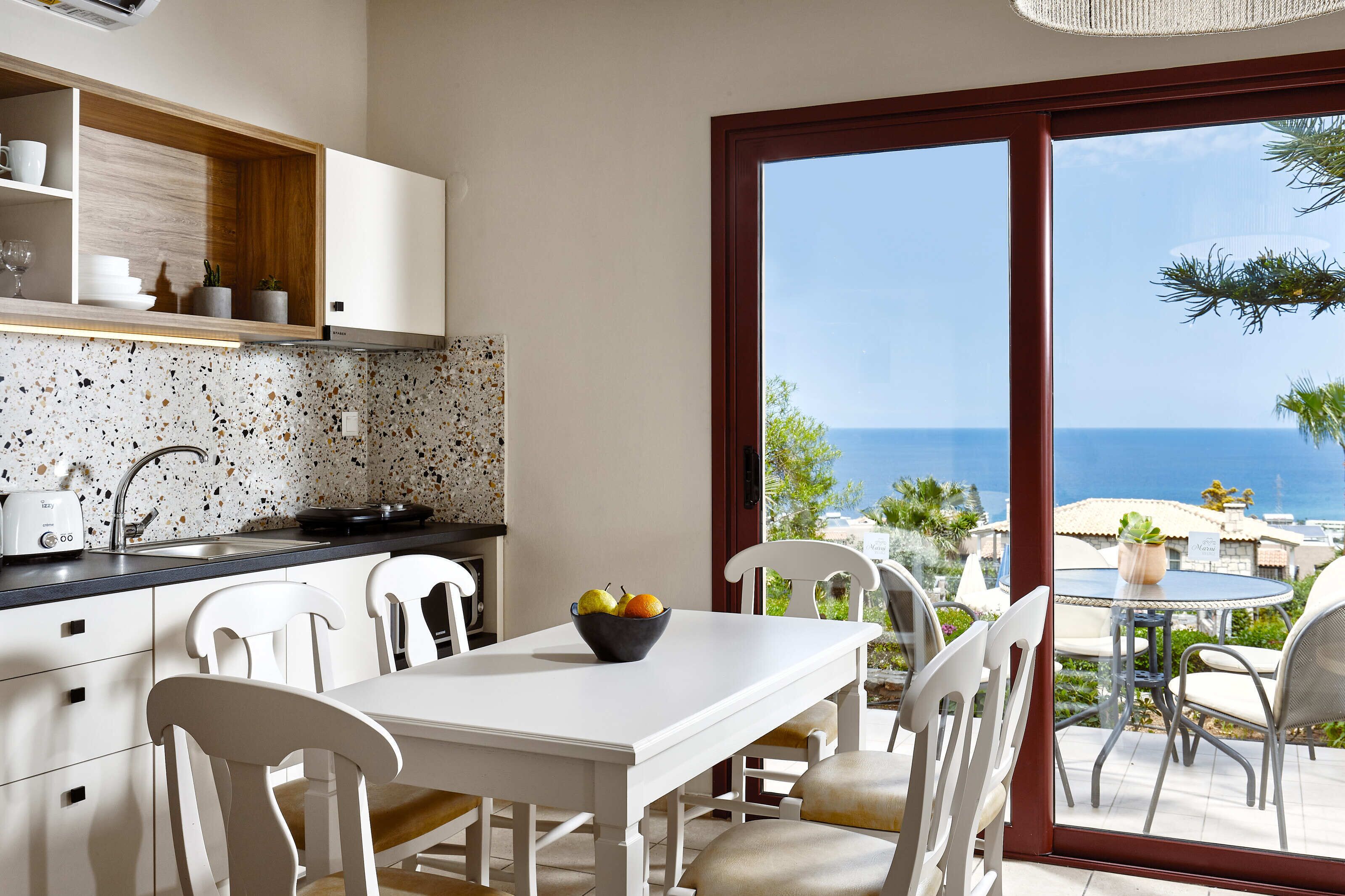 Een moderne keuken met witte kastjes, een spatborden met stippen en een eethoek met een tafel en stoelen. Op tafel staat een fruitschaal, en de kamer heeft een grote schuifdeur die uitkomt op een terras met buitenmeubels.