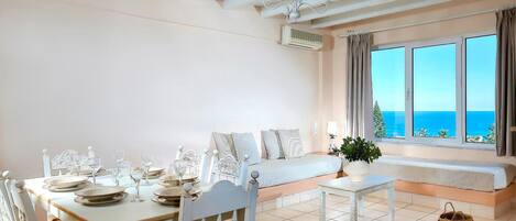 A room with a bed, a dining table set with plates and glasses, a bench, and a view of the sea.