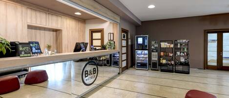 Zona de recepción de hotel con mostrador de madera, un cartel de B&B, una máquina expendedora y un área de descanso con sillas rojas.