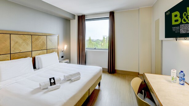 A hotel room with a large bed, a desk, a chair, a window with curtains, and a B&B sign on the wall. - B&B Hotel Padova (Padova)