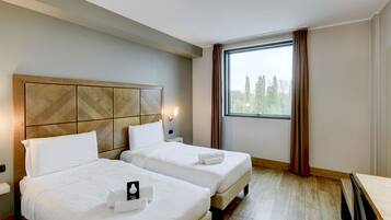 Camera d'albergo con due letti, un'ampia finestra con tende, una testiera in legno e un comodino con una lampada.