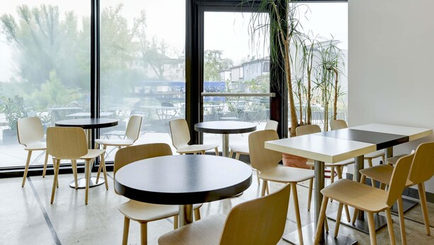 Moderni sisätilojen oleskelualue, jossa on pyöreitä ja suorakulmaisia pöytiä, puisia tuoleja ja suuret ikkunat, joista on näkymä ulos.
