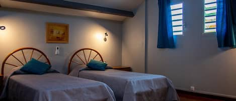 Une chambre avec deux lits, des têtes de lit en bois, un climatiseur fixé au mur, un tableau encadré et une fenêtre avec des rideaux.