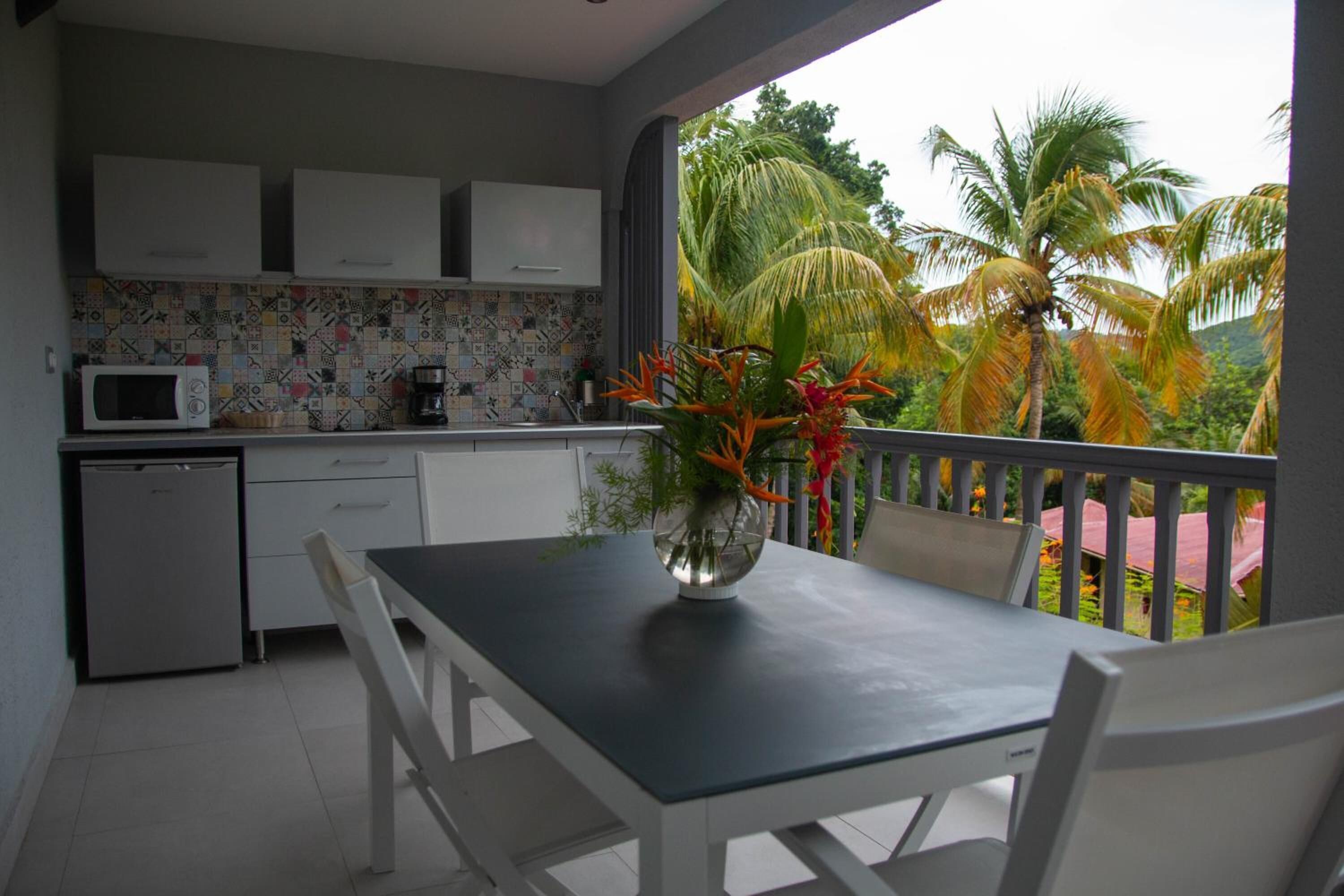 A compact kitchen with white cabinets, a microwave, and a small refrigerator. A dining table with chairs is positioned near a window overlooking palm trees.
