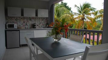 A compact kitchen with white cabinets, a microwave, and a small refrigerator. A dining table with chairs is positioned near a window overlooking palm trees.