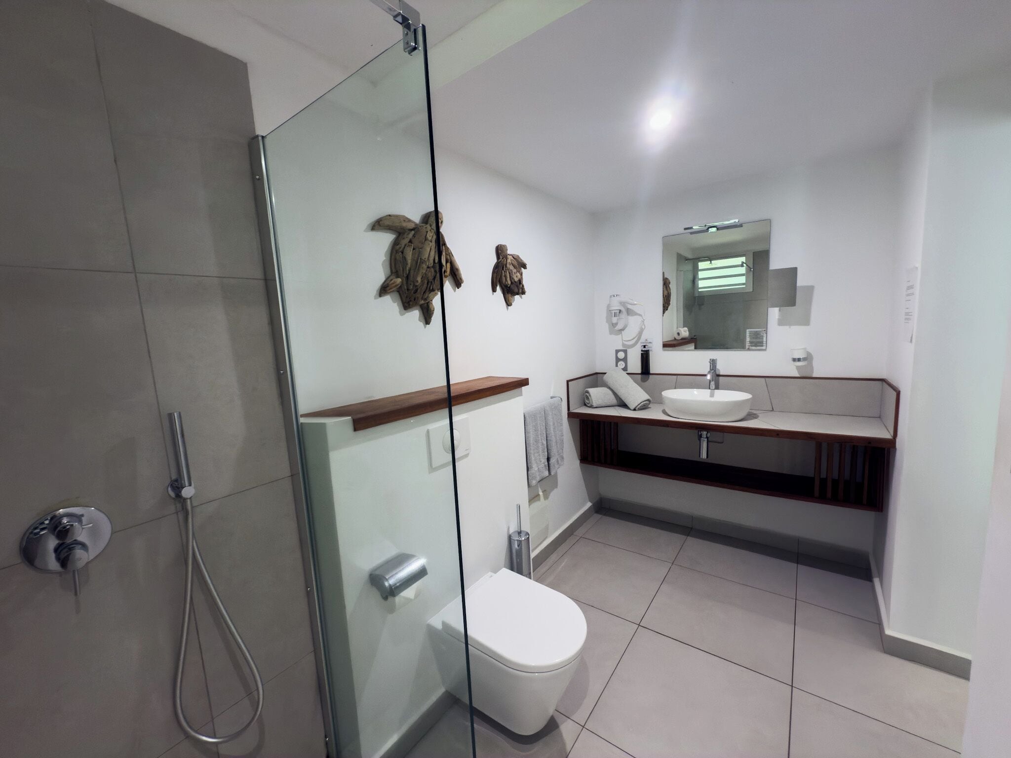 A modern bathroom with a glass shower enclosure, a white toilet, a sink with a mirror above it, and a wooden shelf.