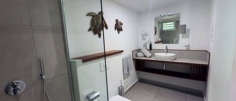 A modern bathroom with a glass shower enclosure, a white toilet, a sink with a mirror above it, and a wooden shelf.