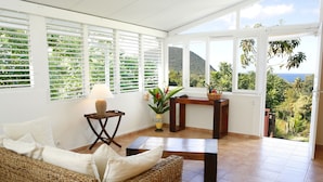 A room with a wicker sofa, a small table with a lamp, and a view of lush greenery and mountains.