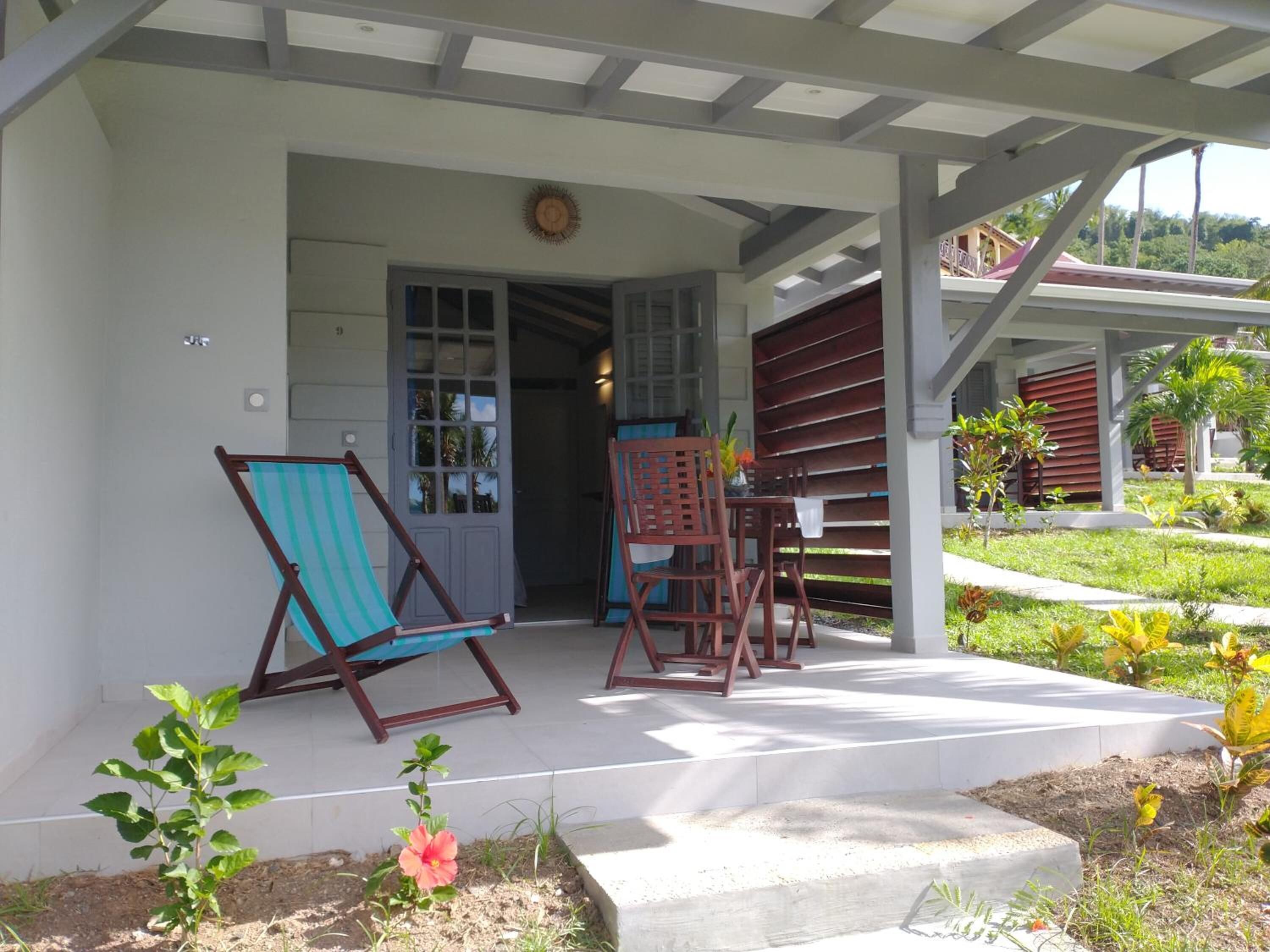 A covered porch with two wooden chairs and a table, a door with glass panels, and a small garden area with plants and flowers.