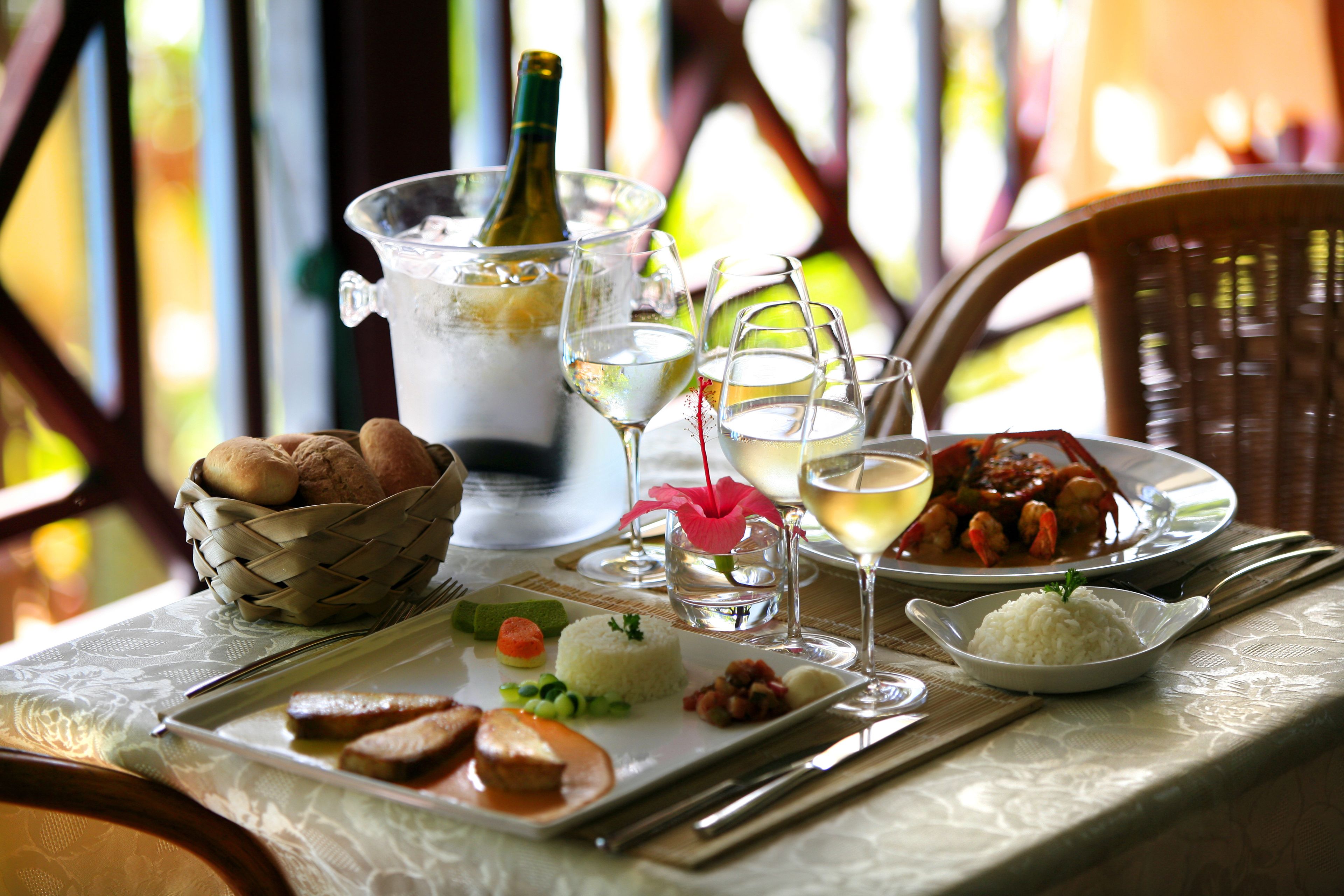 Una mesa dispuesta con una botella de vino blanco, copas, una cesta de pan y platos de comida que incluyen arroz, verduras y un plato de mariscos.