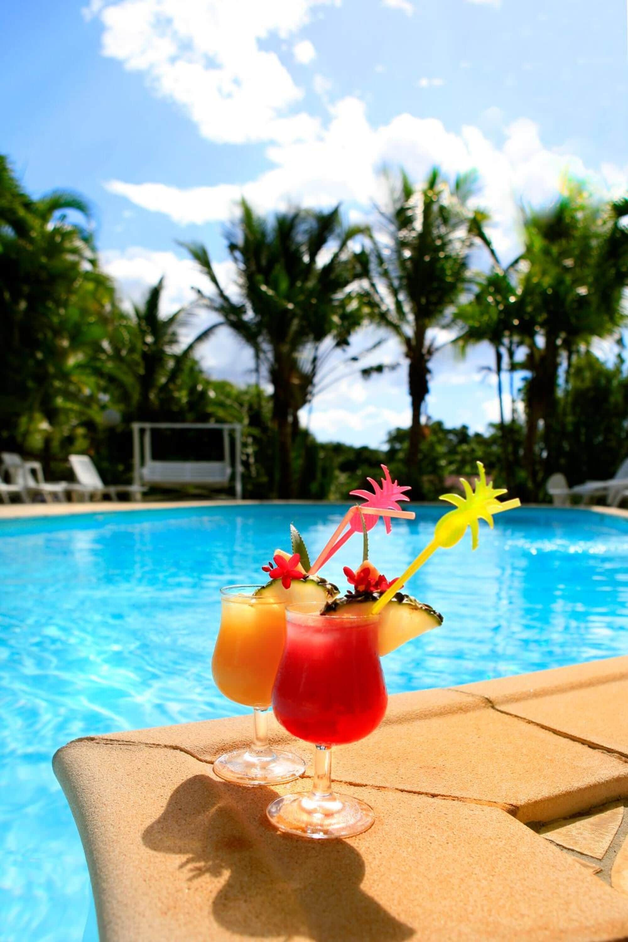 Vista de la piscina con tres bebidas sobre una mesa, rodeada de palmeras y un cielo despejado.