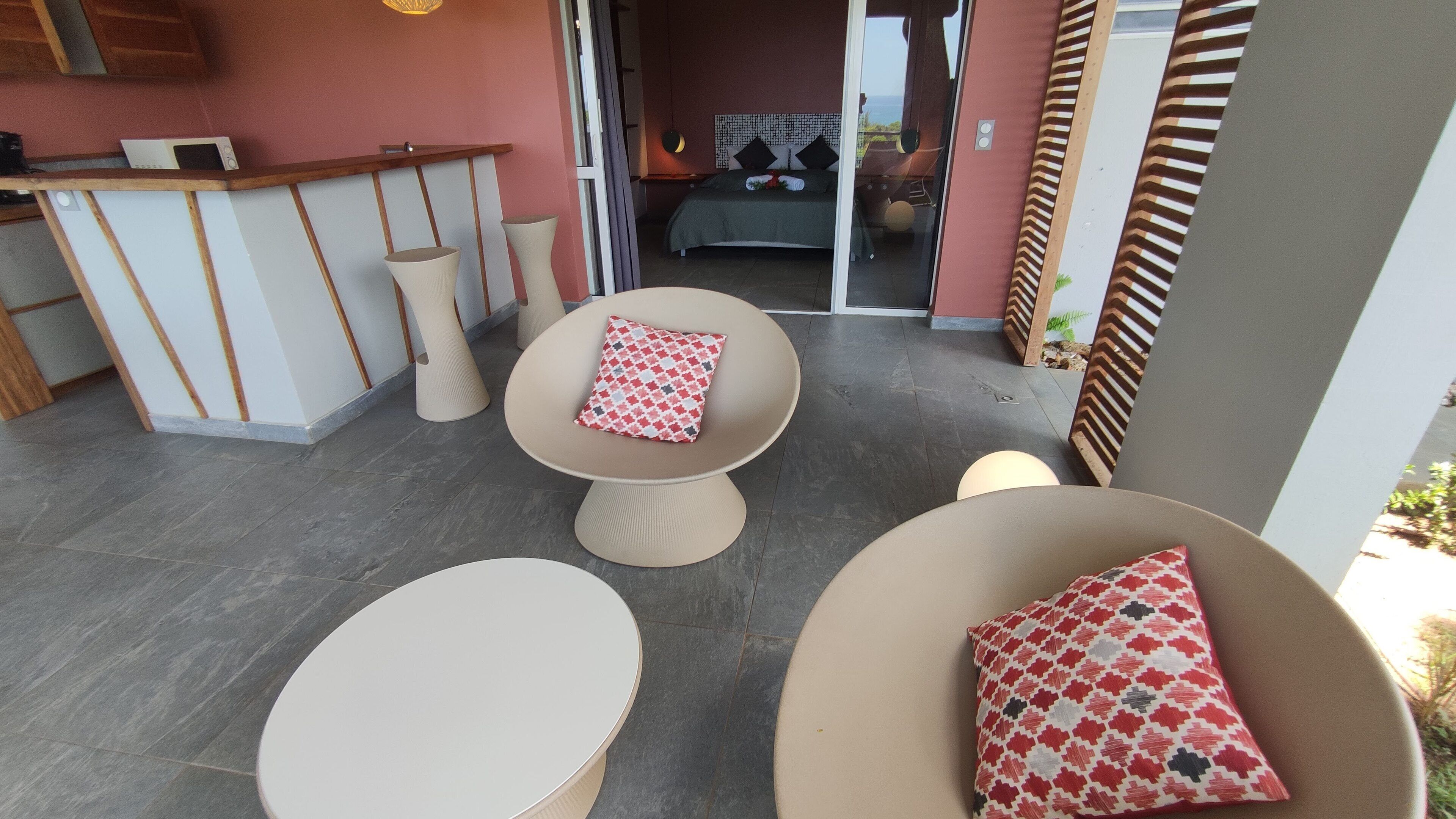 Un espacio moderno al aire libre con dos mesas redondas y sillas, un sofá con un cojín estampado, y vista a un dormitorio con cama y mesita de noche.