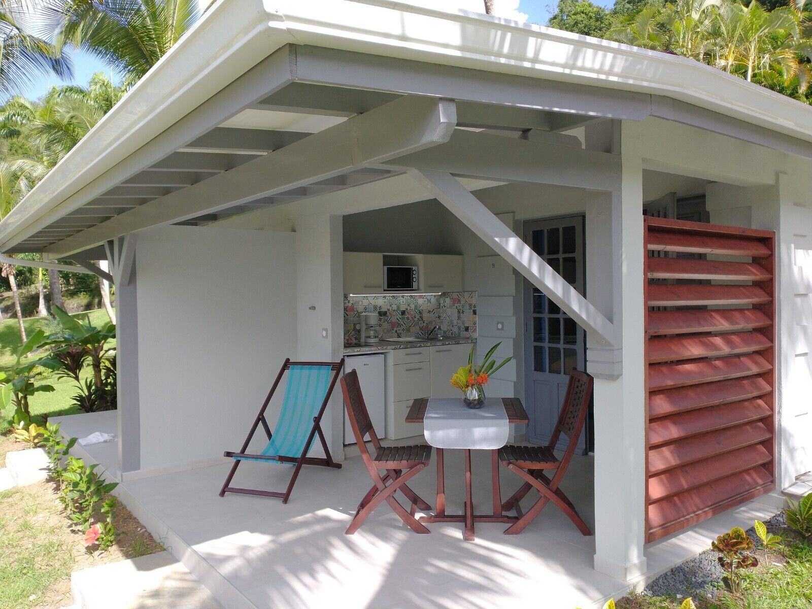 A small, white, covered outdoor area with a kitchen, a table, two chairs, and a blue-striped beach chair.