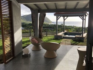 A covered outdoor seating area with modern chairs and a small pool, overlooking a lush landscape and the sea.