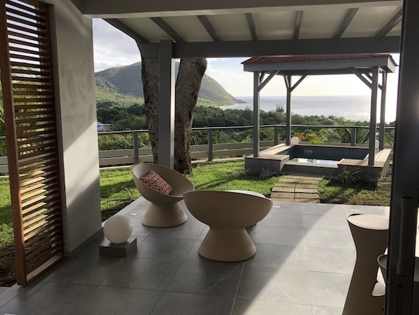 A covered outdoor seating area with modern chairs and a small pool, overlooking a lush landscape and the sea.