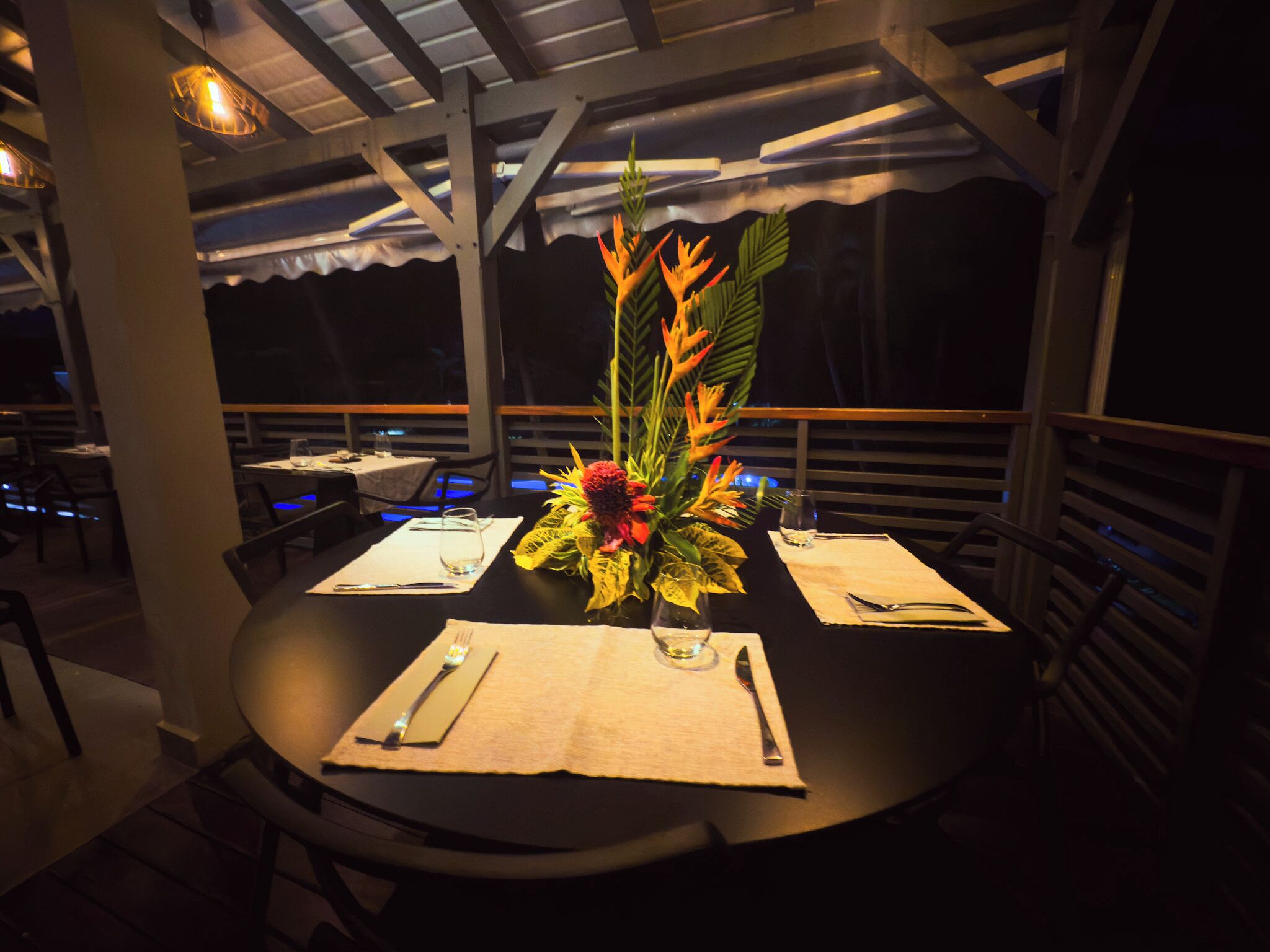 A table set for dining with a centerpiece, under a covered outdoor area with chairs and tables.