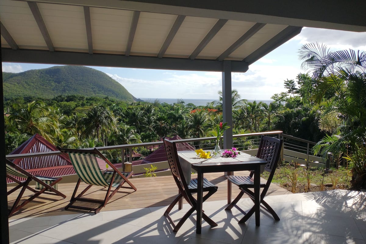 A covered outdoor seating area with a table and chairs, overlooking a lush landscape with mountains and a view of the sea.