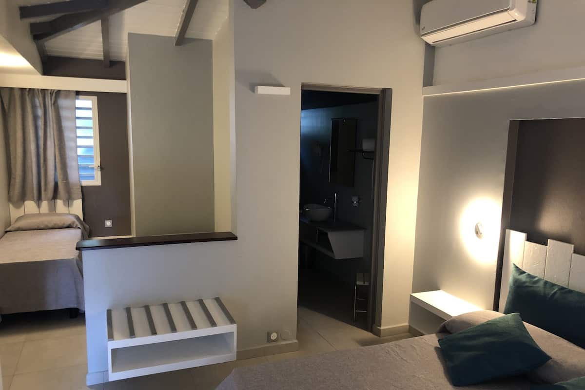A modern hotel room with a bed, a bedside table, a built-in bench, a small seating area, and a bathroom visible through an open door.
