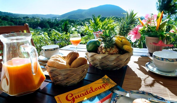 Una mesa de desayuno con una jarra de jugo de naranja, una cesta de pan, un bol de fruta y un folleto turístico.