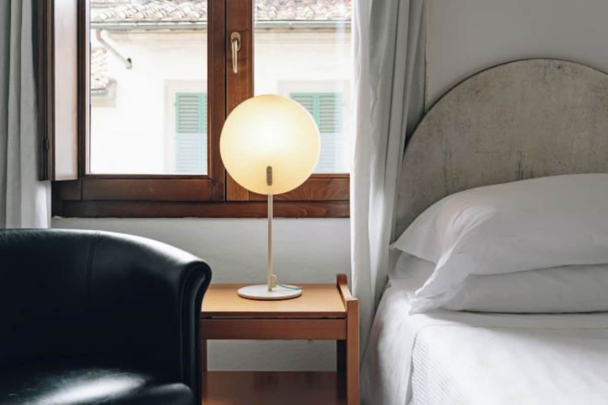 Un dormitorio con cama, mesita de noche con lámpara, una silla y ventana con vistas a edificios.