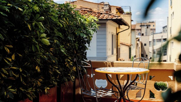 Une petite terrasse extérieure avec une table ronde et des chaises en métal, entourée de verdure et donnant sur un quartier résidentiel.