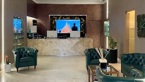 Un hall d’entrée moderne avec un comptoir de réception en marbre, deux canapés en cuir vert, un téléviseur à écran plat diffusant des images d’un paysage urbain et une caméra de surveillance fixée au plafond.