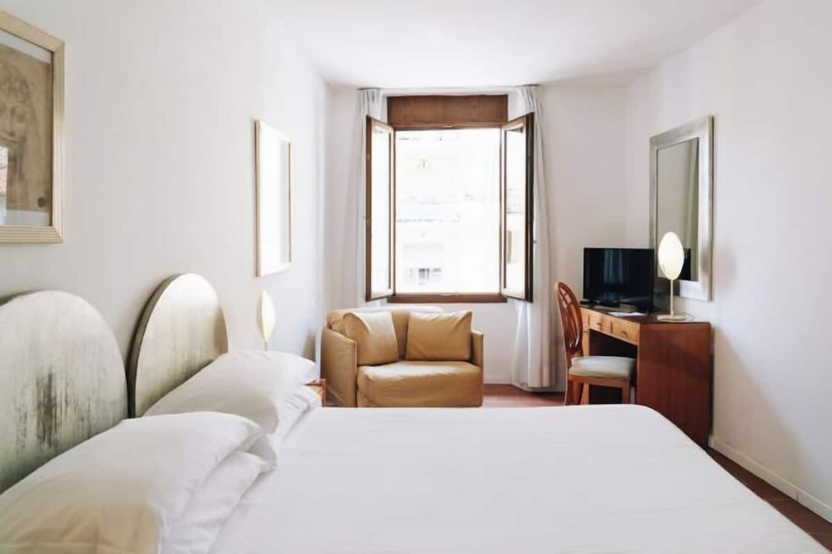 Ein Hotelzimmer mit Bett, Schreibtisch, Sessel, Fernseher und einem Fenster mit Vorhängen.