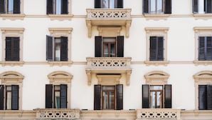 Un immeuble à étages doté d’une architecture symétrique, caractérisé par des fenêtres régulièrement espacées et munies de volets, des balcons avec des garde-corps décoratifs et une façade au design uniforme.