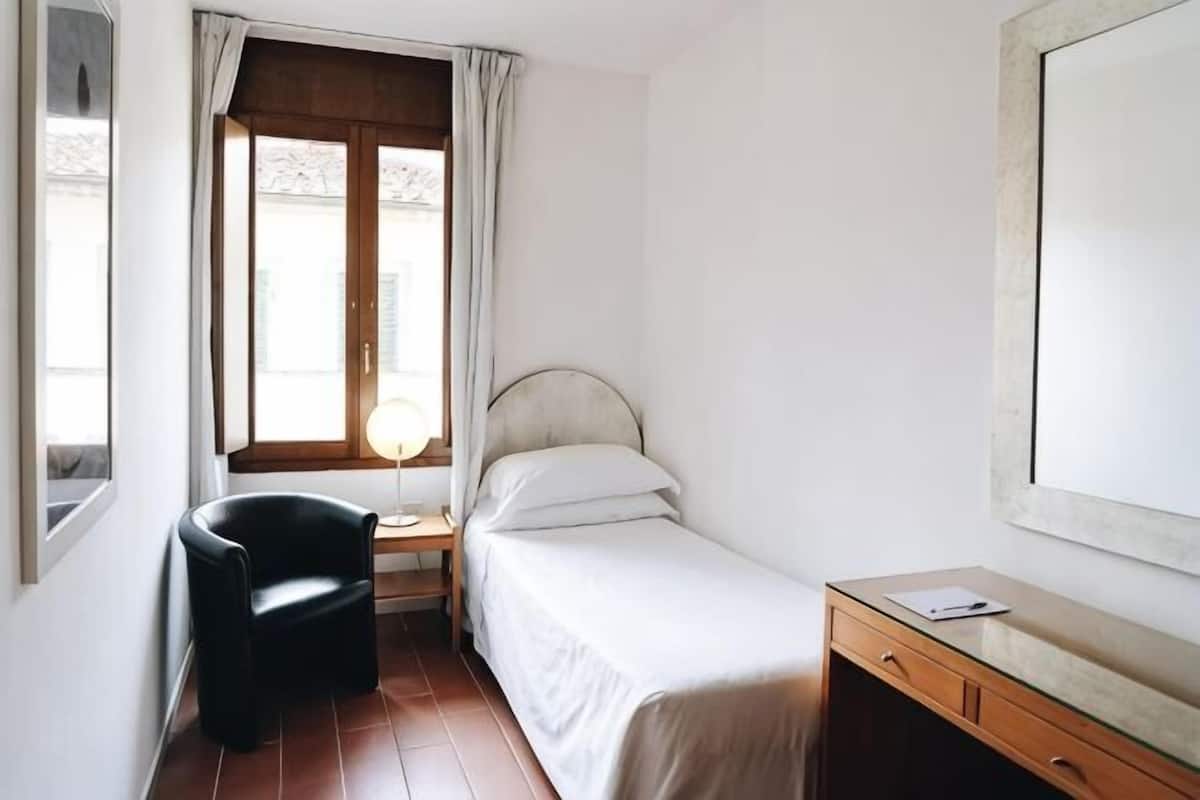 Ein kleiner, sauberer Raum mit Bett, Stuhl, Schreibtisch, einem Fenster mit Vorhängen und einem Spiegel an der Wand.