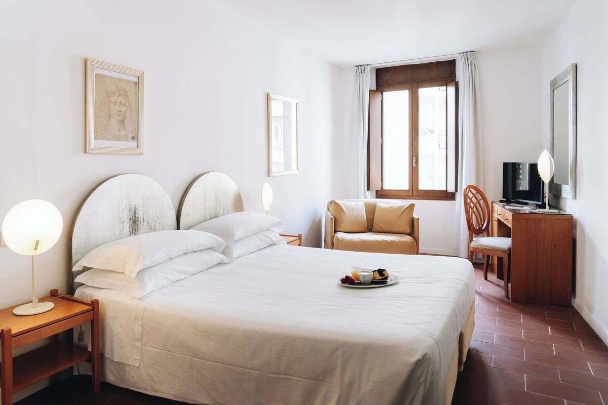 Ein Hotelzimmer mit einem großen Bett, Nachttischen, einem Schreibtisch, einem Sessel und einem Fernseher.
