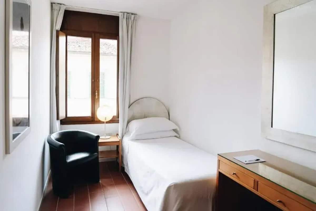 Ein kleiner, sauberer Raum mit einem Einzelbett, einem Sessel, einem Schreibtisch und einem Fenster mit Vorhängen.