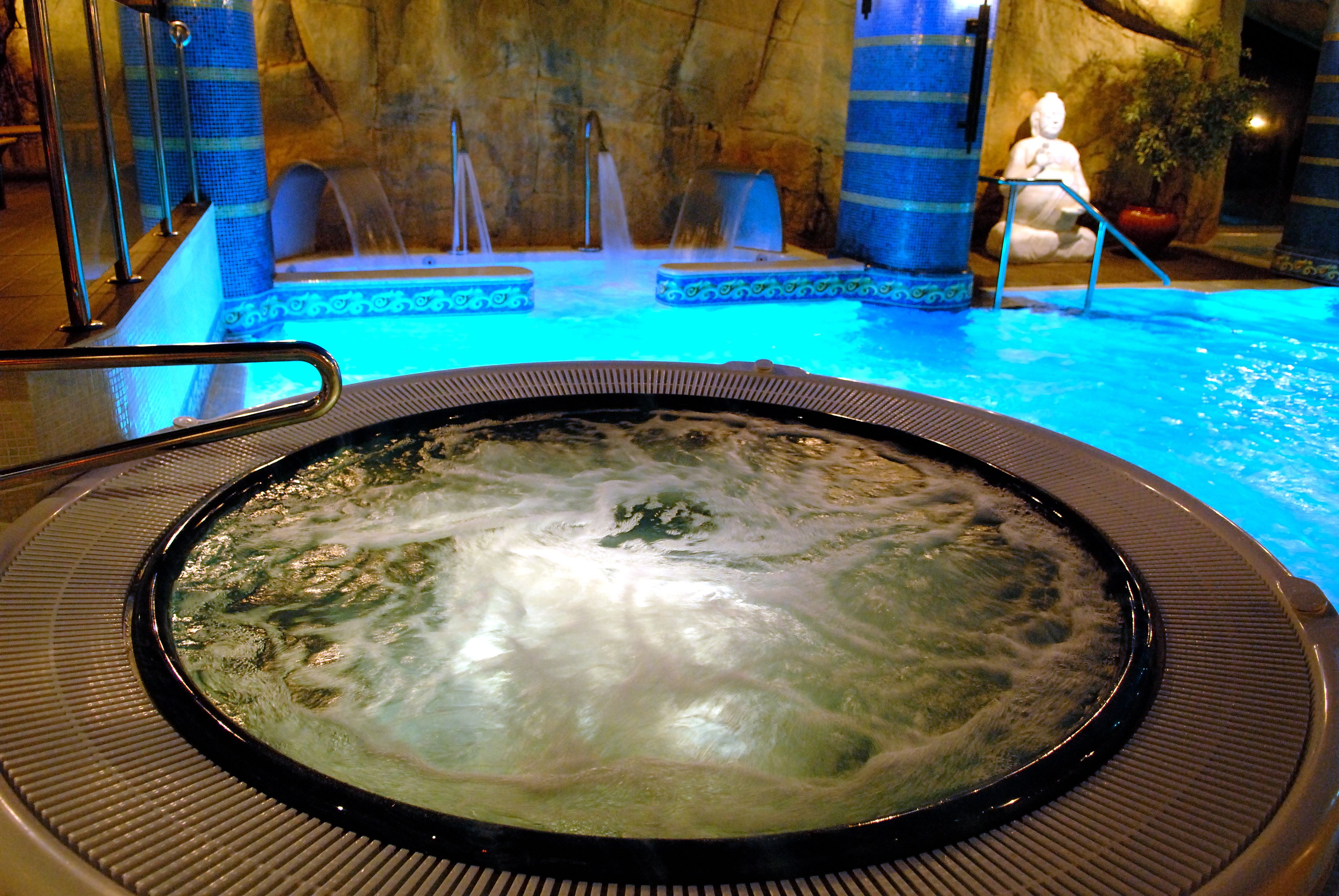 A hot tub with a stone wall backdrop, a statue, and a blue-lit pool area.