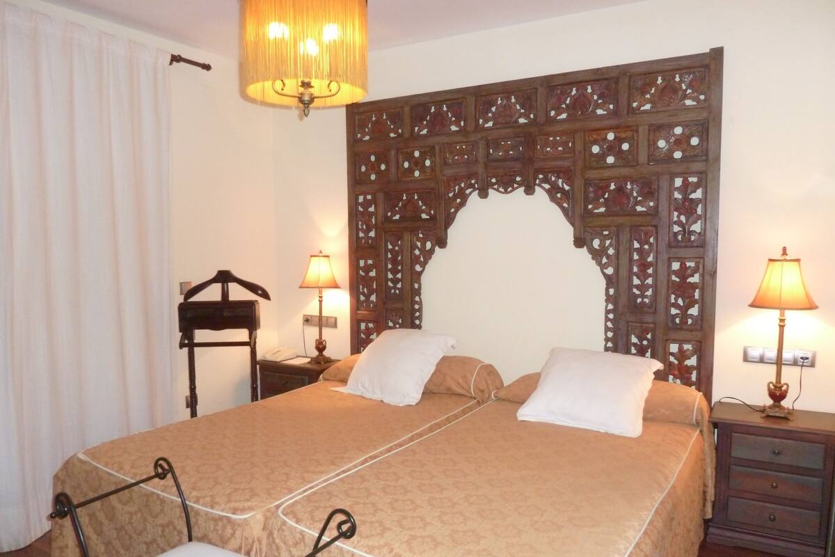 Habitación de hotel ordenada con una cama grande, dos lámparas de noche, un cabecero de madera, una mesita de noche pequeña y una silla.