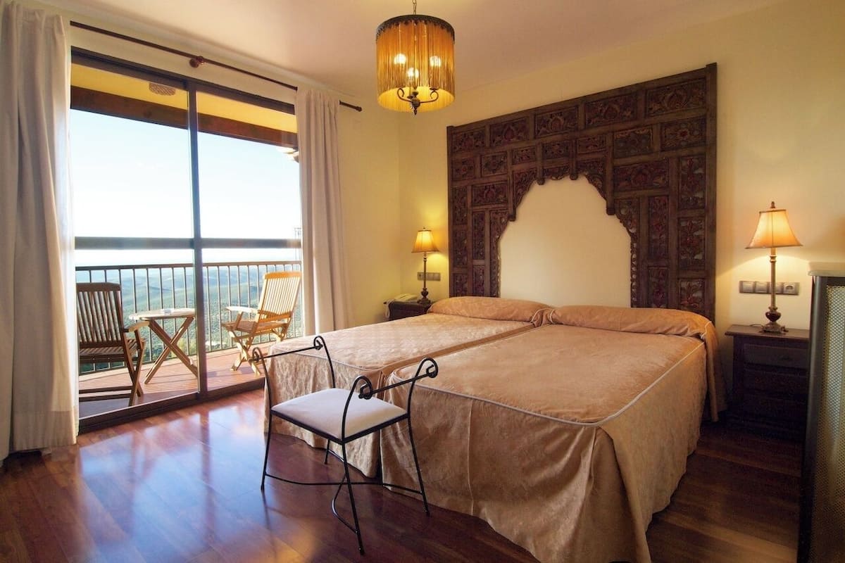 Cama doble con cabecera, dos mesitas de noche con lámparas, una silla y un balcón con sillas.