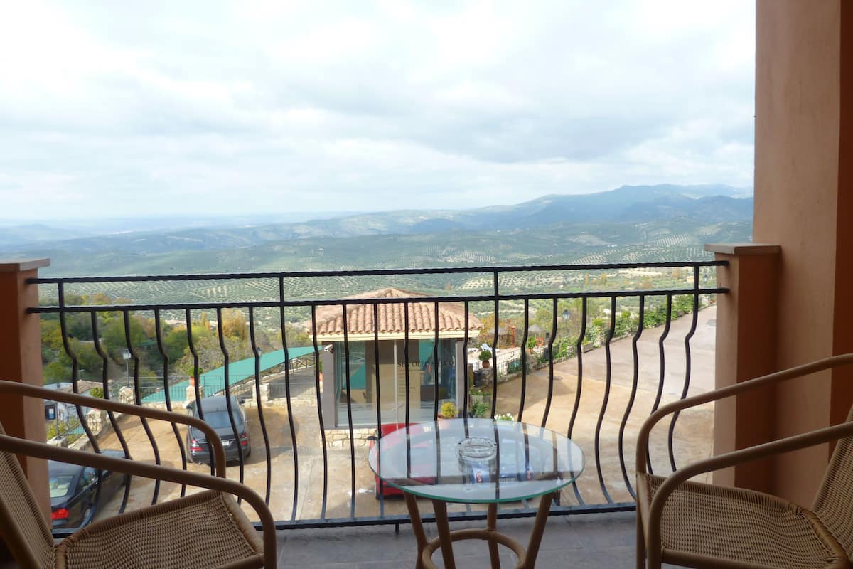 Un balcón con sillas de mimbre y una mesa de vidrio, con vista a un paisaje montañoso con campos en terrazas.
