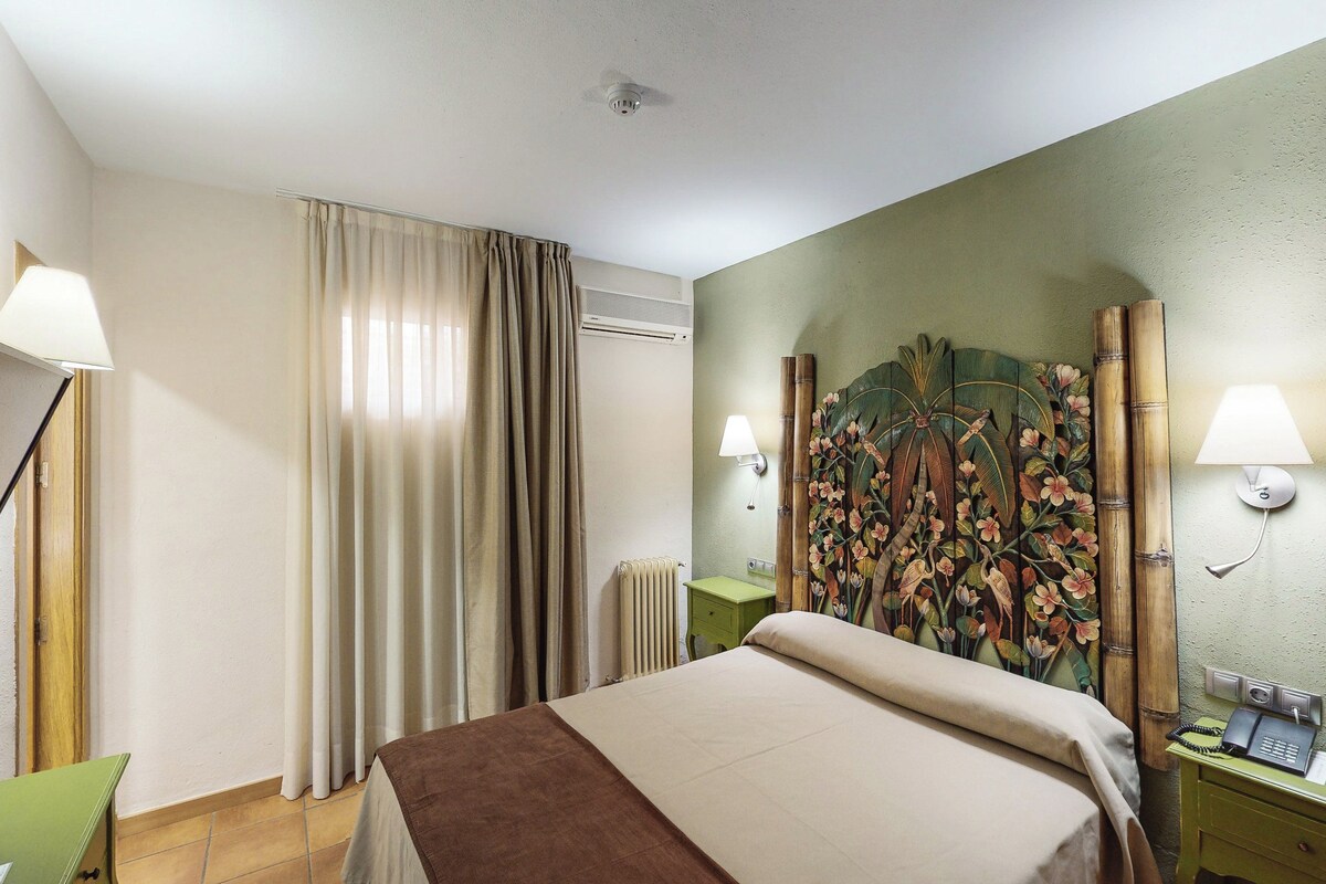 A hotel room with a bed, two bedside lamps, a wooden headboard with a floral design, a radiator, and a green nightstand with a telephone.