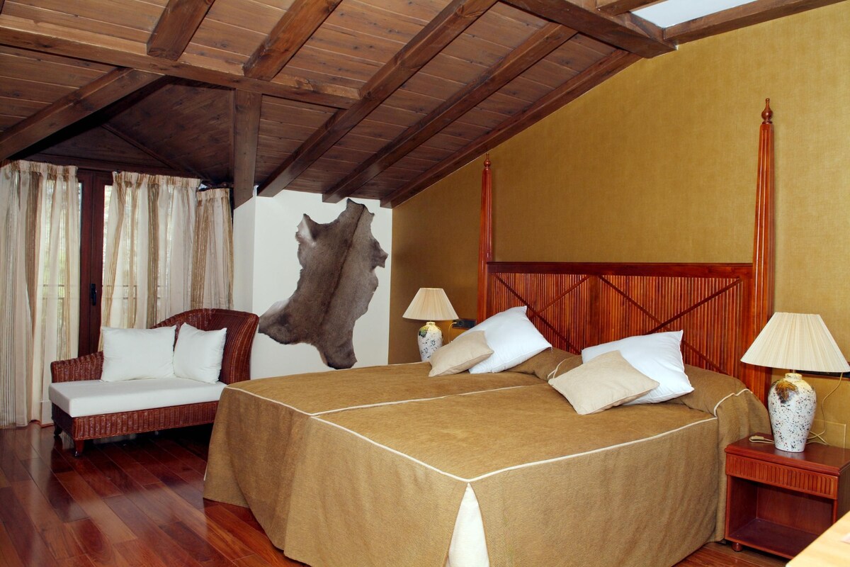 A room with a wooden ceiling, a large bed with a headboard, a sofa, two bedside tables with lamps, and a wall with a large animal hide.