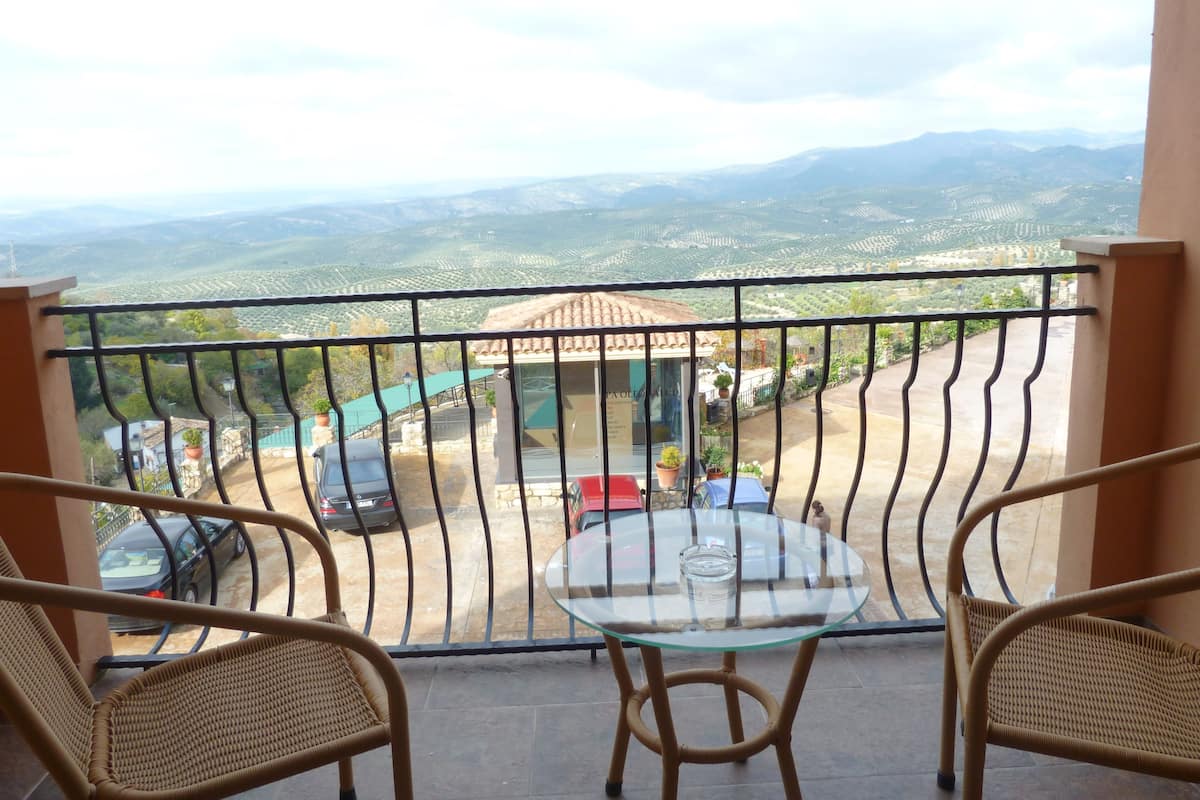 Un balcón con sillas de mimbre y una mesa de vidrio, con vistas a un estacionamiento y montañas a lo lejos.
